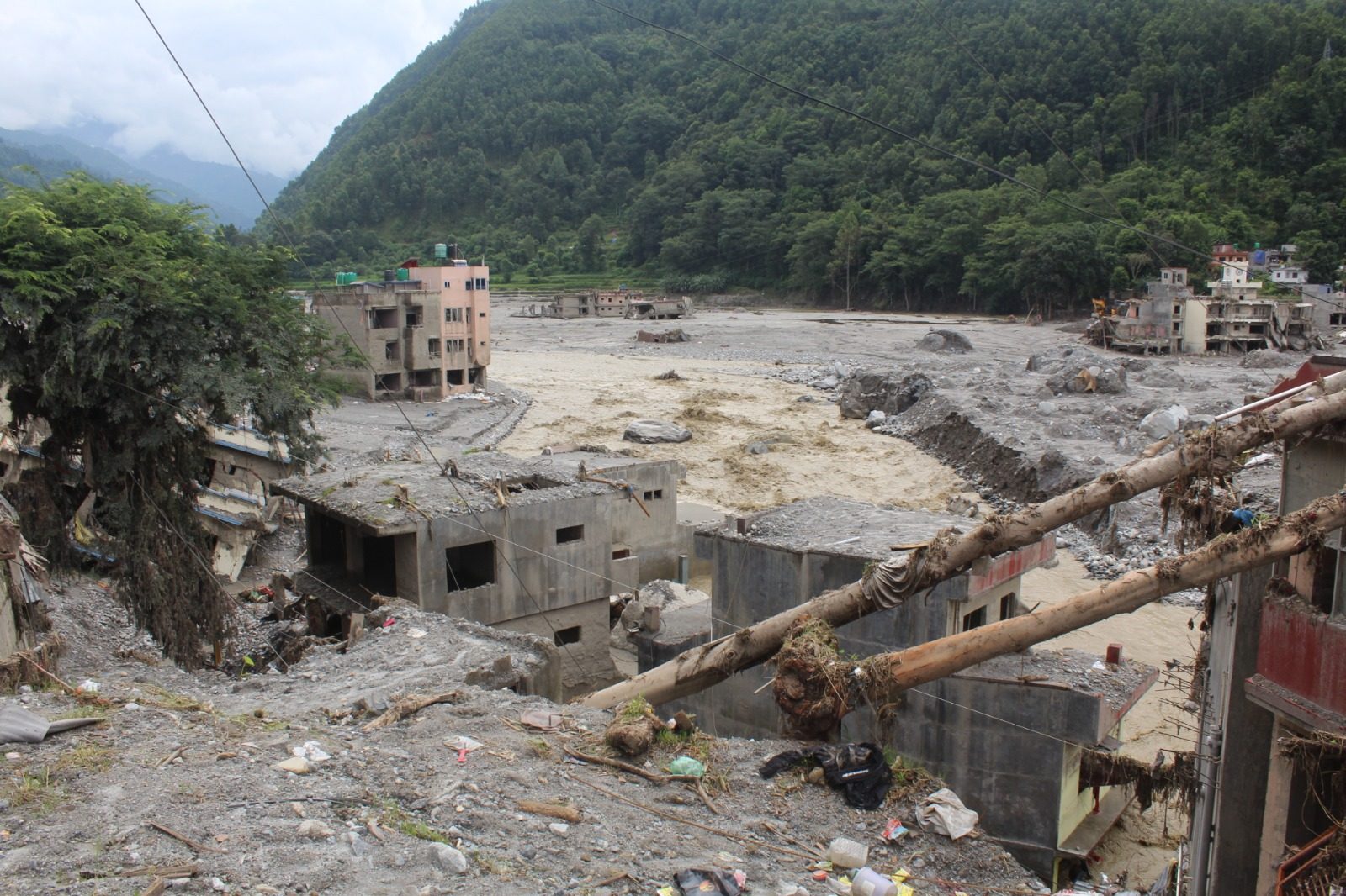 A wide view of a valley town buried in grey silt and boulders, with the river cutting through the middle and a fallen pole in the foreground.