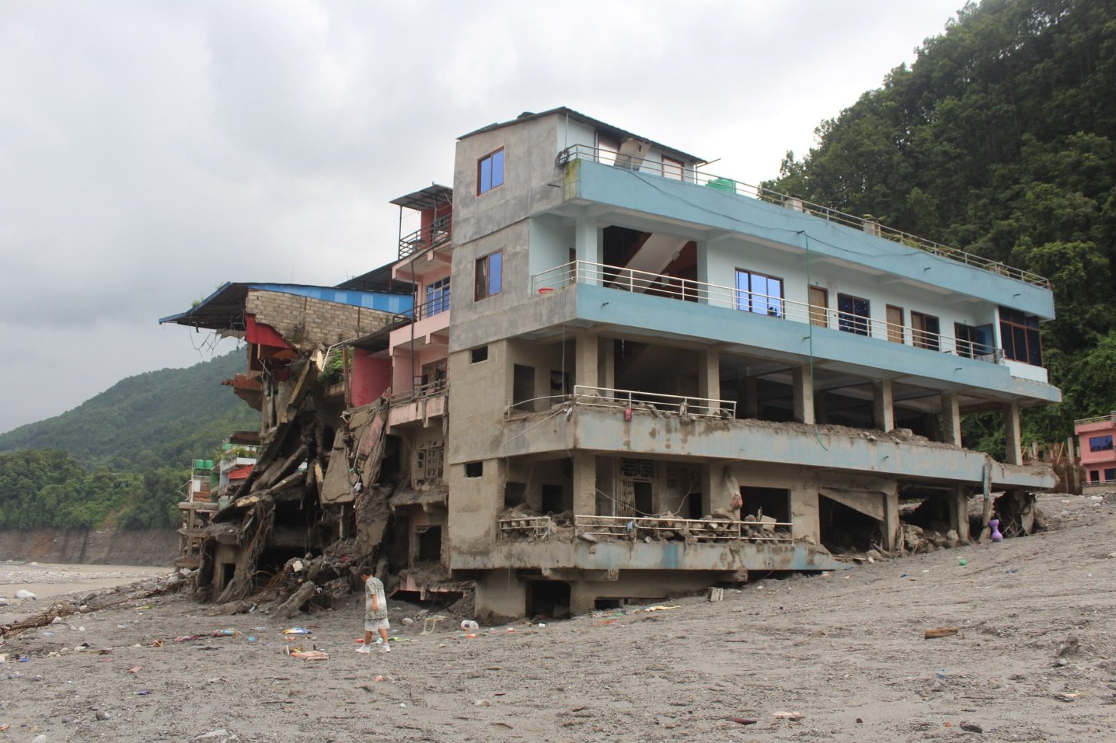 Multi storey buildings left standing on undercut foundations above a bed of grey silt, with one person walking below.